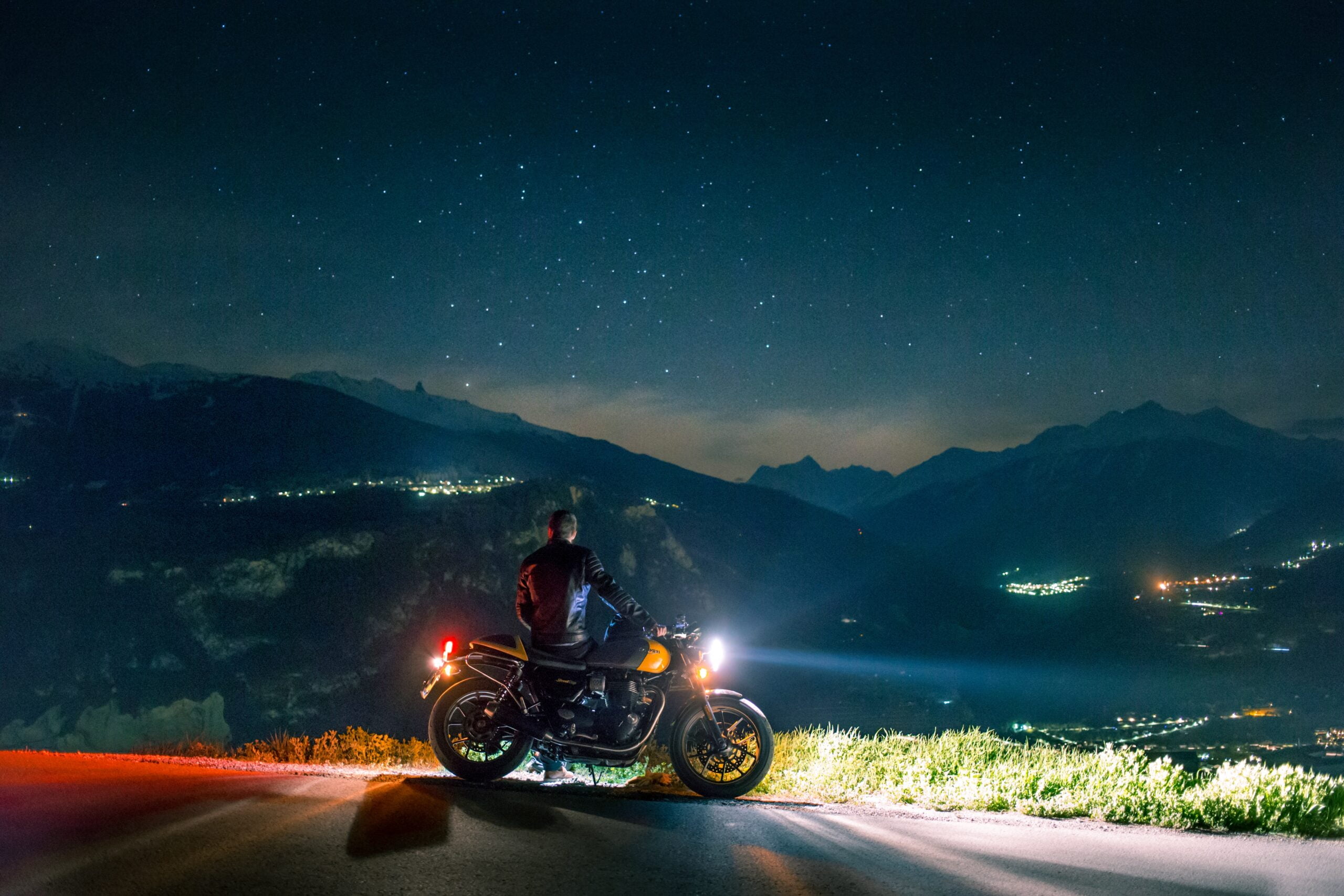 En mann står lent inntil motorsykkelen sin og ser utover en fantastisk fjellandskap på kvelden. Stjernene er fremme og man ser lysene fra små tettsteder.